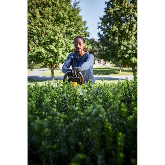 A person in gloves uses a DEWALT hedge trimmer to trim tall green bushes outdoors. Their face is blurred, and trees are