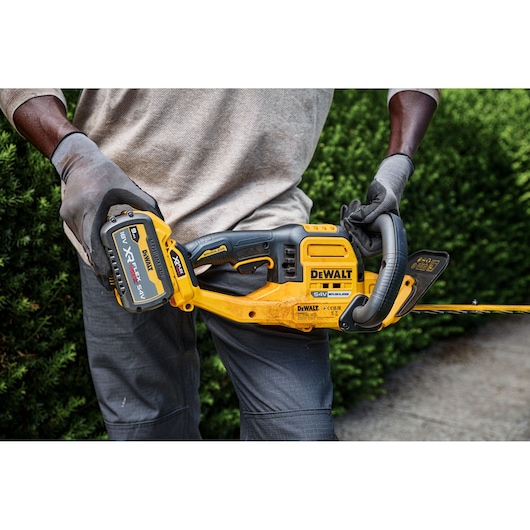 Person holding a DEWALT cordless hedge trimmer outdoors, wearing gloves and standing near bushes.