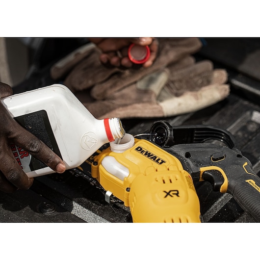 Pouring oil into a DEWALT XR chainsaw with gloves and cap nearby on a black surface.