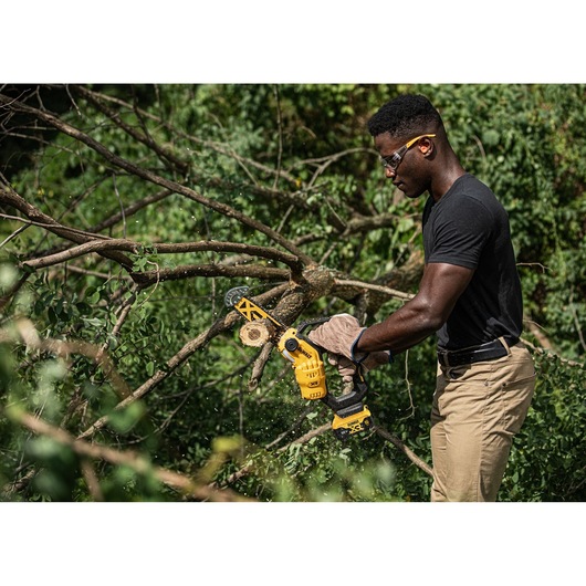 Person uses DEWALT cordless chainsaw to cut tree branches outdoors.