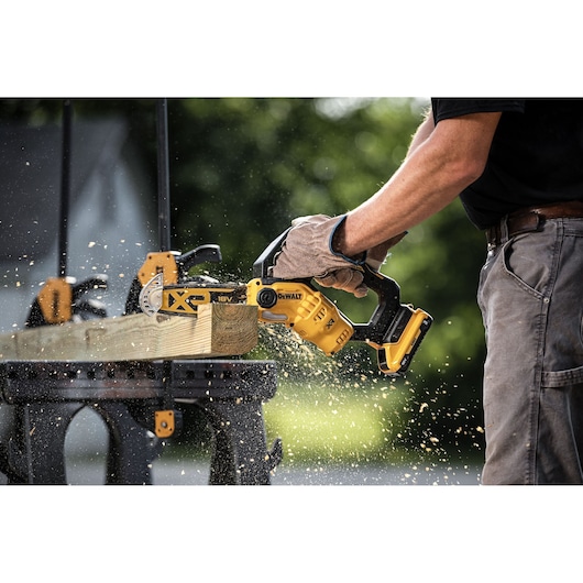 Person using a DEWALT cordless chainsaw to cut wood, with sawdust flying and work gloves worn.