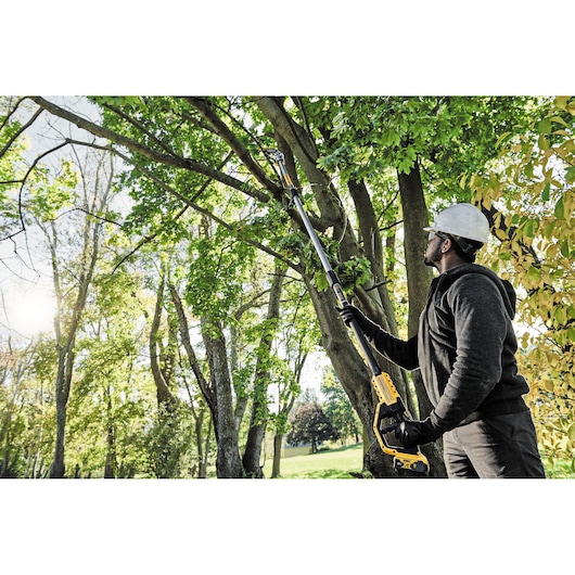 A person wearing protective gloves and a hard hat is using a DEWALT cordless pole saw (model DCMPS567P1) to trim branches