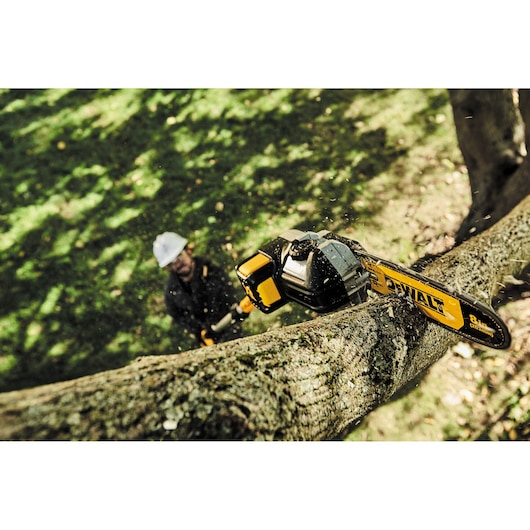 A DEWALT cordless pole saw is being used to cut a thick tree branch outdoors. The person operating the tool is blurred in