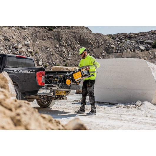 A worker in high-visibility clothing is unloading a DEWALT DCMPW1600 pressure washer from the back of a pickup truck in a