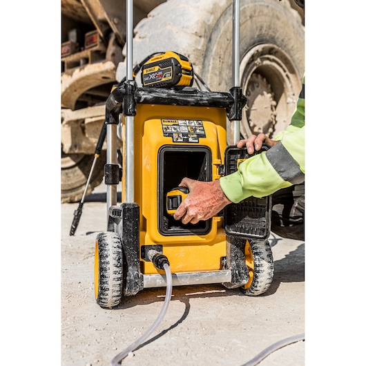 A person is inserting a battery into a yellow DEWALT portable power washer (model DCMPW1600) outdoors on a dusty