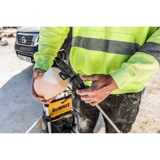 A person wearing a high-visibility yellow jacket is holding a soap or detergent bottle and connecting it to a DEWALT
