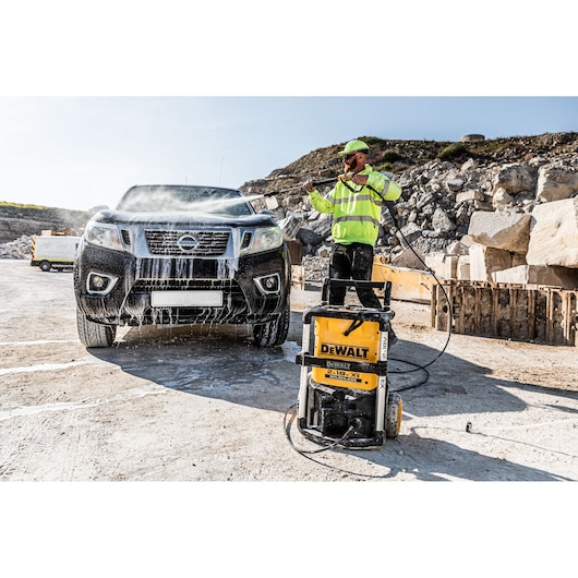 A worker uses a DEWALT pressure washer (model DCMPW1600) to clean a black pickup truck outdoors at a construction site. The