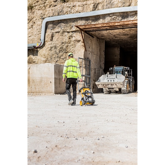 A worker wearing a high-visibility jacket is pulling a DEWALT portable pressure washer (model DCMPW1600) across a dusty