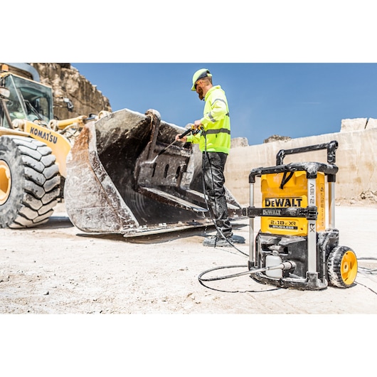 A person in safety gear is using a DEWALT portable pressure washer (model DCMPW1600) to clean the bucket of a construction