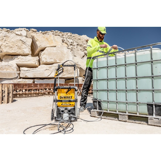 A DEWALT pressure washer (model DCMPW1600) is positioned on a sandy outdoor worksite next to a large water storage tank. A