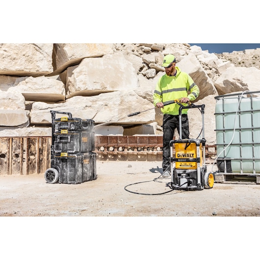 A person wearing a high-visibility jacket operates a DEWALT DCMPW1600 portable pressure washer outdoors, cleaning stacked