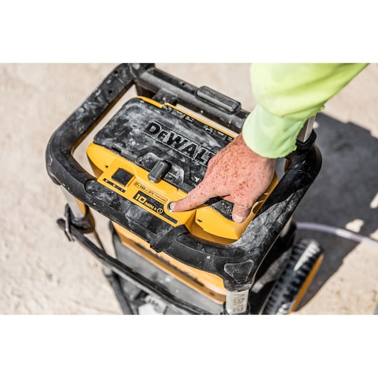 A close-up of a person pressing a button on the DEWALT DCMPW1600 portable pressure washer, which is dusty and used outdoors