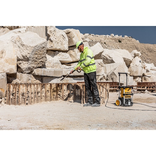 A worker wearing a high-visibility jacket and helmet uses a DEWALT pressure washer outdoors in a rocky industrial setting