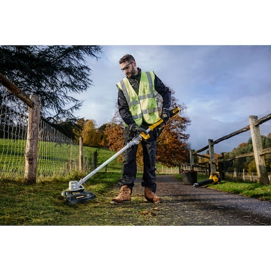 A person wearing a reflective safety vest and work boots is using a DEWALT cordless grass trimmer (model DCMST561N) to trim grass along a fence in an outdoor park or rural area.