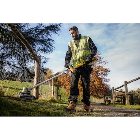 A person wearing a high-visibility vest uses a DEWALT cordless string trimmer (model DCMST561N) to trim grass along a wire fence in an outdoor rural setting.