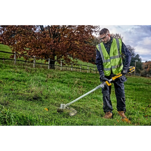 A person wearing a high-visibility vest is using a DEWALT cordless grass trimmer (model DCMST561N) outdoors on a grassy area, with trees and a wooden fence in the background.