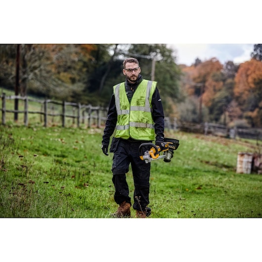 A person wearing a reflective safety vest and work gloves is walking outdoors on a grassy field, holding a DEWALT DCMST561N cordless grass trimmer. There is a wooden fence and trees in the background.