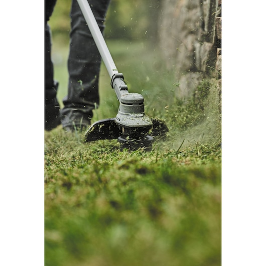 Close-up of a DEWALT cordless string trimmer being used to cut grass near a stone wall. Grass clippings are visible flying