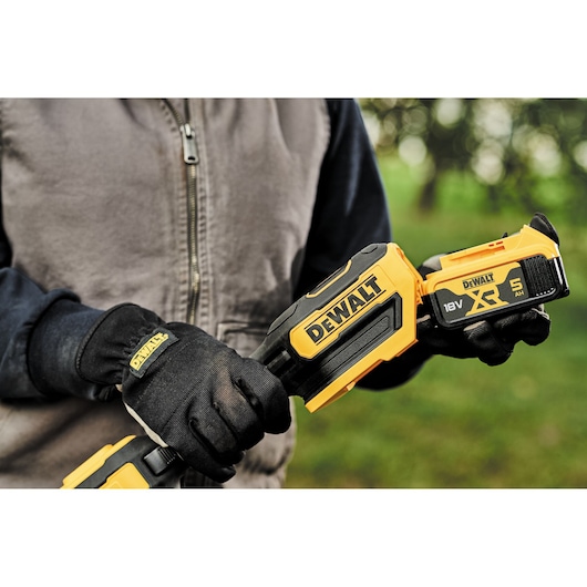 Close-up of a person wearing black gloves and a grey jacket, holding a DEWALT 18V XR cordless tool with a 5Ah battery
