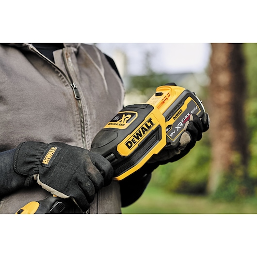 A person wearing gloves and a jacket is holding a DEWALT cordless brushless power tool with an 18V XR battery in an outdoor