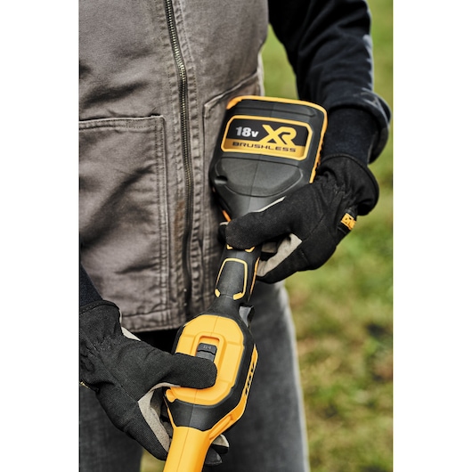 Close-up of a person wearing work gloves holding a DEWALT 18V XR Brushless power tool outdoors.