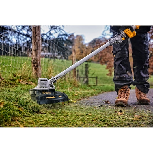 A person using a DEWALT battery-powered brushless grass trimmer to cut grass along a fence outdoors. The trimmer is being