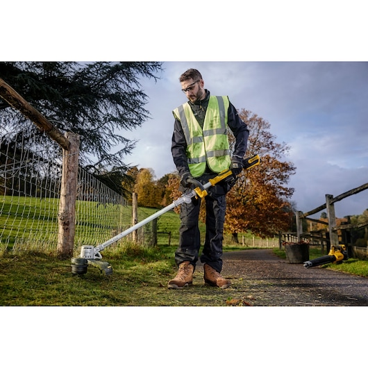 A person wearing a high-visibility vest and work gloves uses a DEWALT cordless string trimmer (DCMST561P1) to trim grass