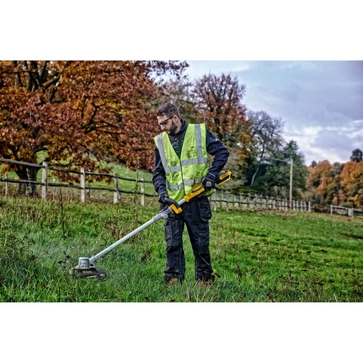 A person wearing a high-visibility vest and gloves uses a DEWALT DCMST561P1 cordless string trimmer to cut grass in a field