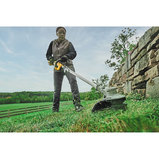 A person using a DEWALT cordless string trimmer outdoors to cut grass along a stone wall, with a green field and fence in