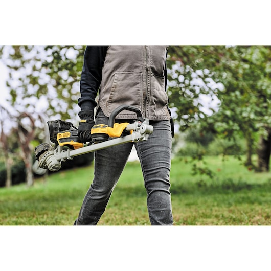 A person outdoors holding a DEWALT cordless string trimmer, model DCMST561P1, designed for lawn and garden maintenance.