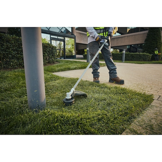 Person using DEWALT cordless grass trimmer near sidewalk and lamp post in landscaped outdoor area.