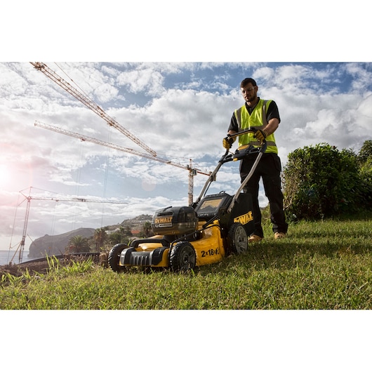 A person wearing a yellow safety vest operates a DEWALT DCMW564N lawn mower on a grassy area outdoors. Construction cranes and a partly cloudy sky are visible in the background.