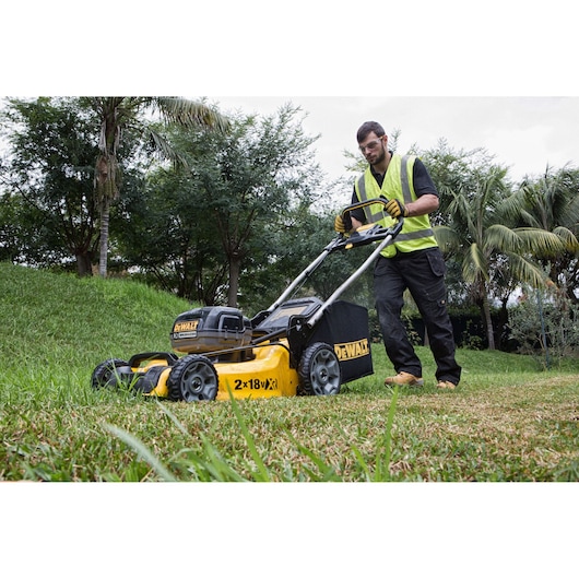 A person wearing a safety vest is using a DEWALT cordless lawn mower outdoors on a grassy area, surrounded by trees and plants.