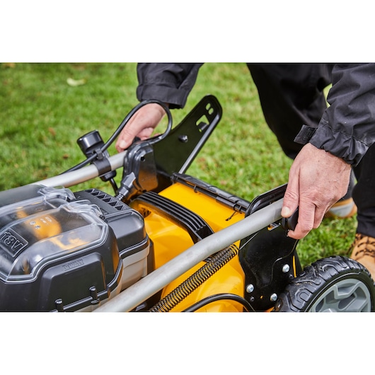 Close-up of a person adjusting the handle of a yellow DEWALT cordless lawn mower on grass.