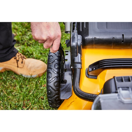 Close-up image showing a person adjusting the wheel height lever on a yellow DEWALT lawn mower, while standing on grass.