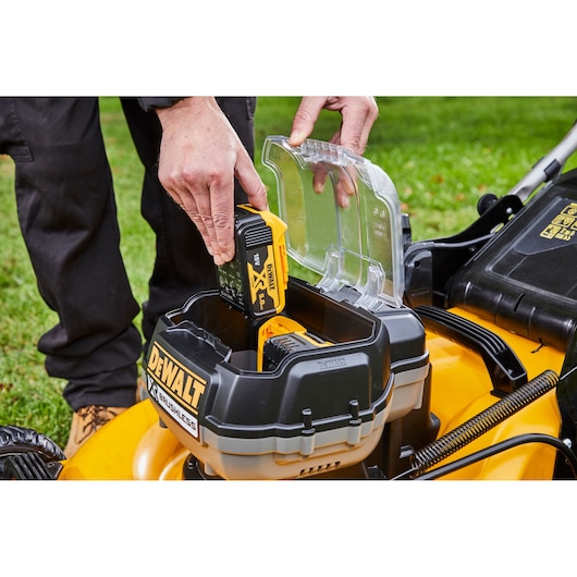 A close-up of a person inserting a DeWalt 18V battery into a yellow and black DeWalt brushless lawn mower, with the battery compartment open and grass visible in the background.