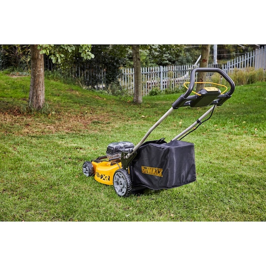 A DEWALT DCMW564N cordless lawn mower is positioned on a grassy lawn, with trees and a fence in the background.