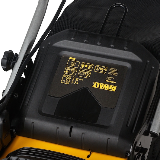 Close-up view of a DEWALT DCMW564N lawn mower featuring the control panel and safety information label with various icons and instructions.