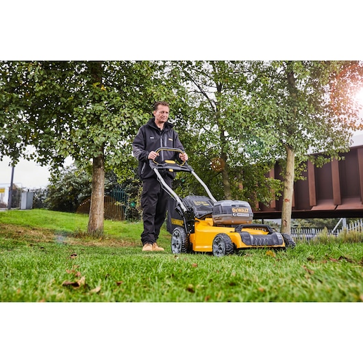 A person using a DEWALT DCMW564P2 cordless lawn mower outdoors on green grass, with trees and a building in the background.