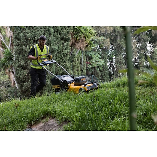 A person wearing a high-visibility vest operates a DEWALT cordless lawn mower on a grassy hillside, surrounded by various