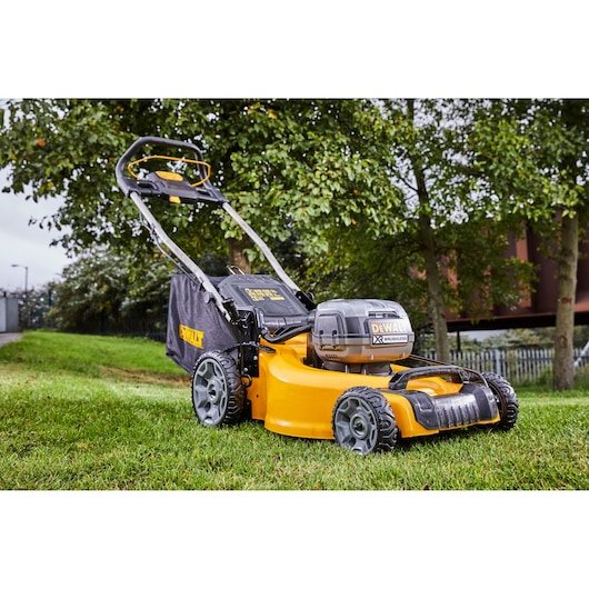 A DEWALT cordless electric lawn mower with large wheels and a grass collection bag, shown outdoors on a lawn with trees in