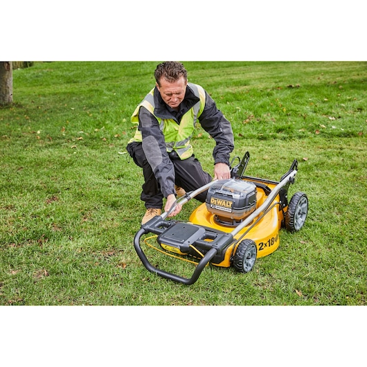 A person wearing a safety vest and work clothes is adjusting a DEWALT DCMW564P2 cordless lawn mower on a grassy area.