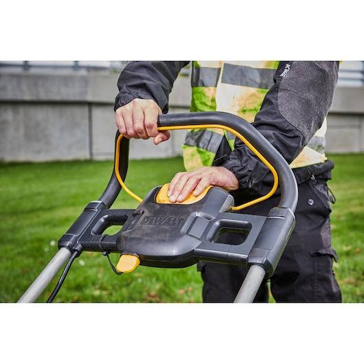 Close-up of a person operating the handle and control panel of a DEWALT cordless lawn mower outdoors, with visible grass and