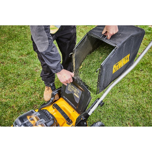 A close-up view of a person emptying the grass collection bag from a DEWALT lawn mower on a grassy lawn.