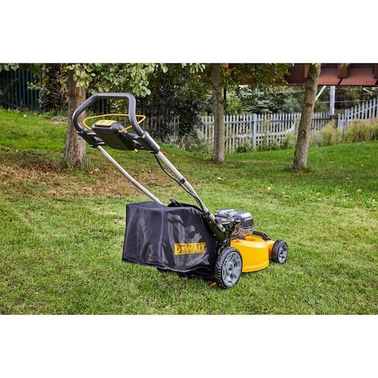 A DEWALT cordless lawn mower with a black grass collection bag and a yellow body, positioned on a grassy lawn near trees and