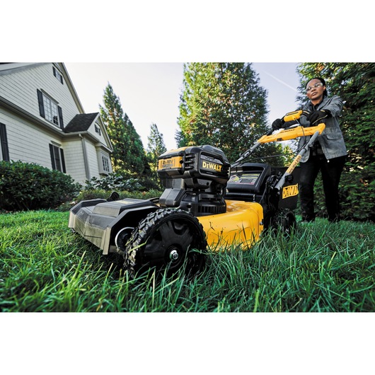 A person is pushing a DEWALT battery-powered lawn mower on a grassy lawn near a house, with trees and bushes in the background. The focus is on the yellow and black mower.