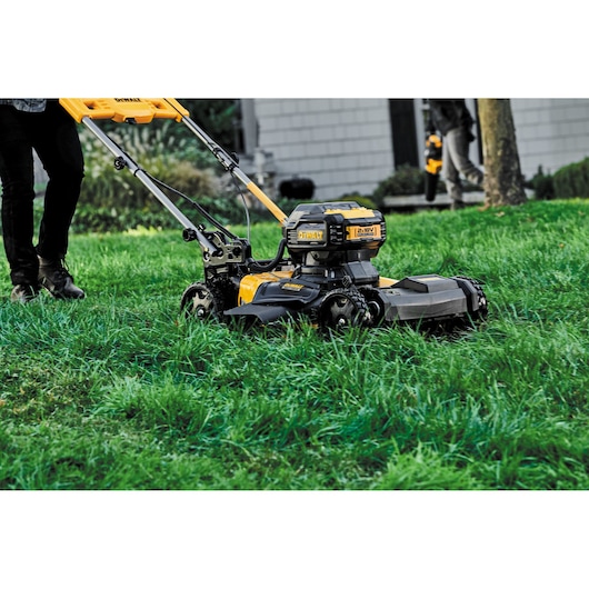 A DEWALT cordless lawn mower being pushed across a grassy lawn outdoors, with a person visible walking behind it.