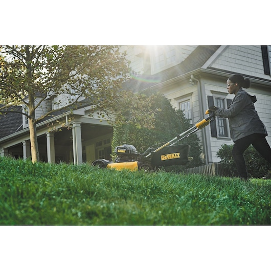 A person is pushing a yellow and black DEWALT lawn mower across a grassy yard in front of a house. The scene is outdoors with sunlight shining over the lawn.