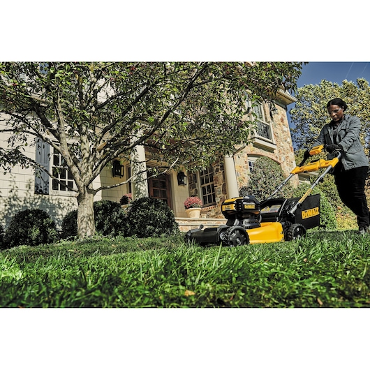 A person is pushing a yellow and black DEWALT lawn mower across a grassy lawn in front of a house, surrounded by green bushes, a tree, and flower pots.