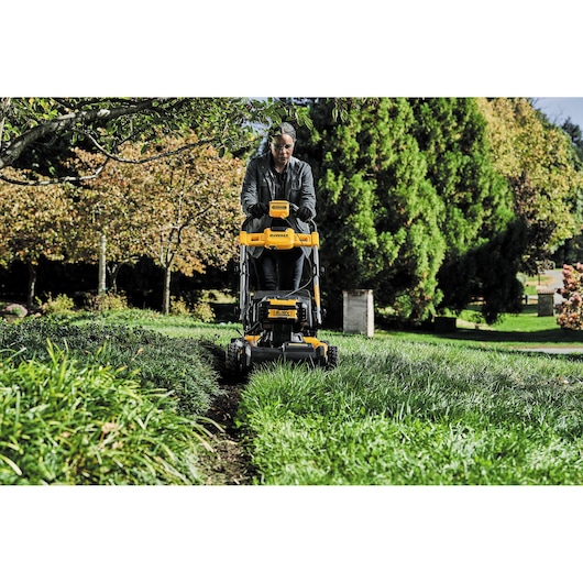 A person using a DEWALT DCMWSP564N lawn mower in a garden surrounded by green grass and trees. The person’s face is blurred for privacy.